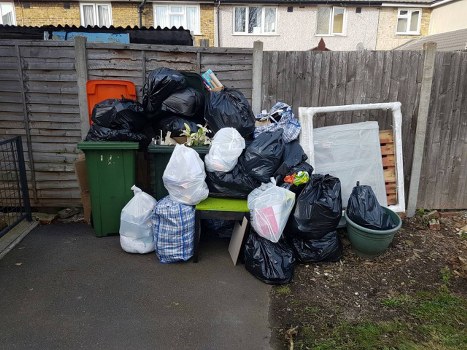 A collection of various waste bags and containers arranged outdoors on a paved surface near a wooden fence. Several black and white rubbish bags are stacked and placed on a green bench and ground, with some bags appearing full and tied closed. A large green wheeled bin is positioned on the left side, and next to it is a smaller green pot with a lid. Behind the waste, a leaning white frame or panel lies against the fence, and a few loose items, such as a small cardboard box or packaging, are visible among the bags. The fence is constructed from vertical wooden panels, and a row of residential buildings can be seen in the background. The scene suggests a typical rubbish collection area for local waste management services, such as Commercial Waste Islington, in an urban setting near the postcode area consistent with central London.