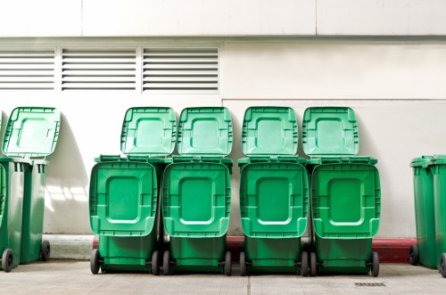 A row of nine identical green wheeled rubbish bins arranged on a concrete pavement next to a plain white wall with horizontal ventilation slats. The bins are made of durable plastic with textured surfaces, and each has a hinged lid in a slightly darker shade of green. They are positioned in a line, with some bins placed directly in front of others, creating a staggered appearance. The environment appears to be an urban external area, possibly at the rear or side of a commercial building in Islington, with natural daylight illuminating the scene. The bins are clean and closed, ready for waste collection, which aligns with the services of a local rubbish removal company like Commercial Waste Islington. The red-painted curb features slight weathering, and the overall setting suggests a practical waste management point close to a building facade, typical for professional rubbish disposal tasks in the London borough.