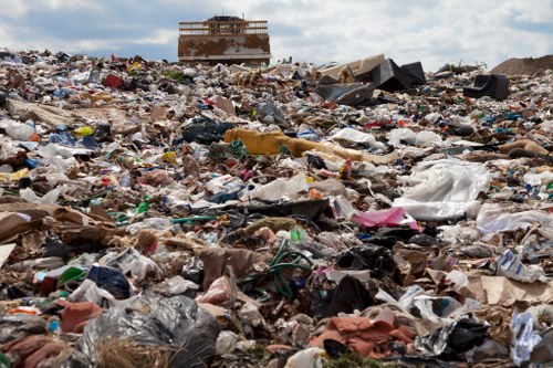 A large, uncontained landfill site with a vast expanse of mixed waste materials, including plastic bags, cardboard, paper, and miscellaneous debris, spread across the ground. The waste appears haphazardly piled, with some items partially crushed or torn, and others loosely scattered. In the background, there is a distant, partially constructed or abandoned building with a rusty, metal frame, set against a cloudy sky. The site is outdoors, likely near an urban area such as Islington, where commercial waste removal services by companies like Commercial Waste Islington may be required. The overall scene highlights a typical industrial or commercial refuse dumping area, emphasizing the scale and variety of waste involved in rubbish collection and disposal activities.
