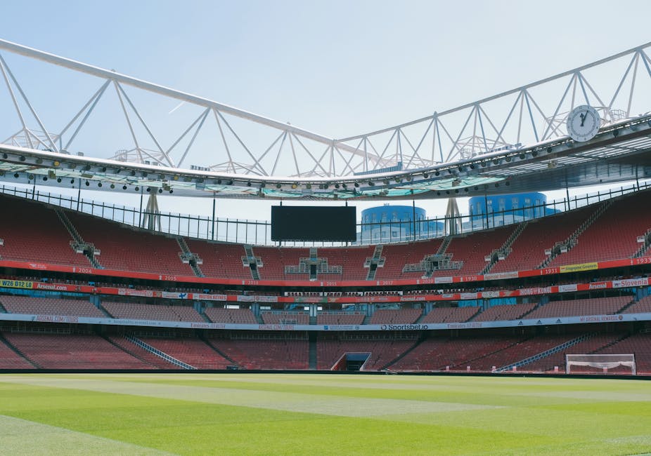 A large, empty stadium with multiple tiered seating sections in red and blue, surrounding a well-maintained grass pitch. The seats are arranged in a circular pattern with some sections having black ba