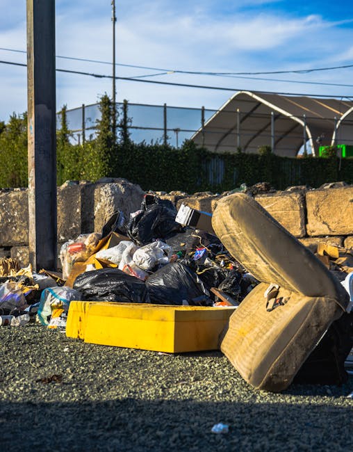 A collection of overflowing rubbish and recycling bins situated on a paved urban street near a parking area. The large central bin is made of grey plastic, with a partially open blue lid, overflowing 